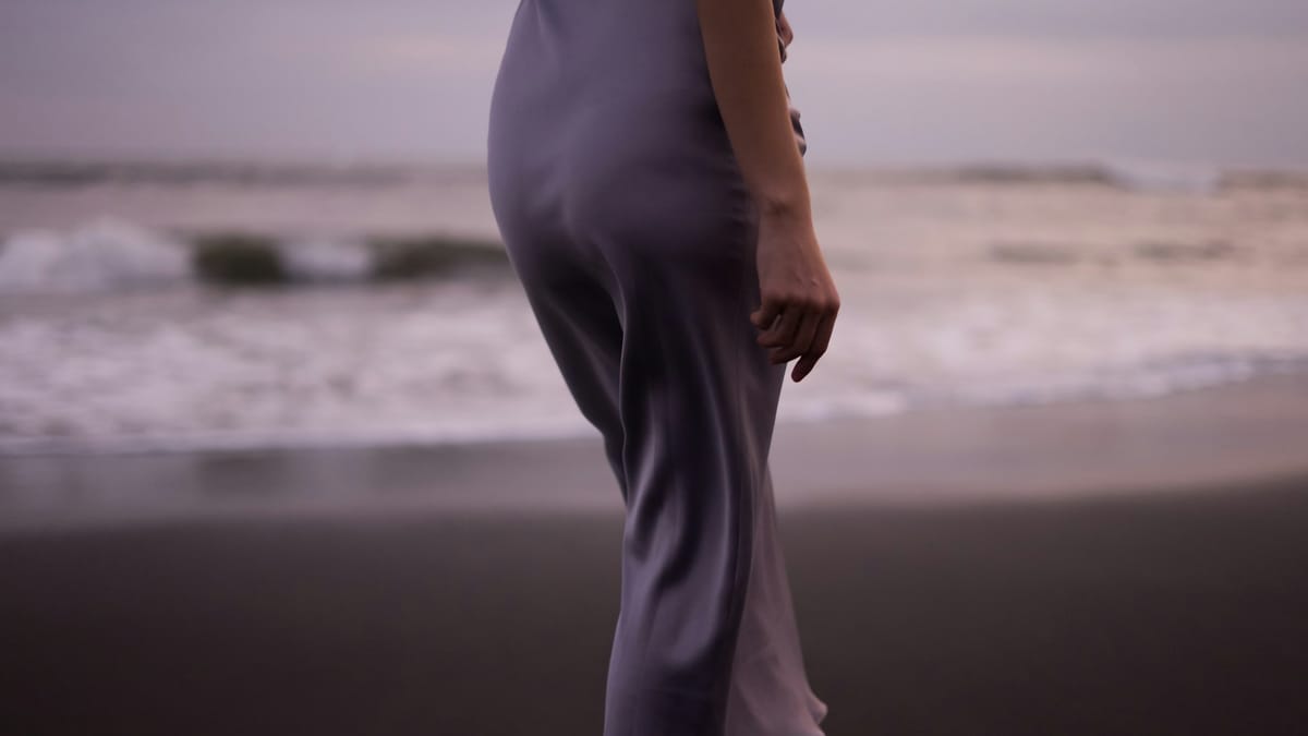 A figure in a slate satin midi walking the shore at dusk, the kind of single garment that reads correctly across multiple formality levels
